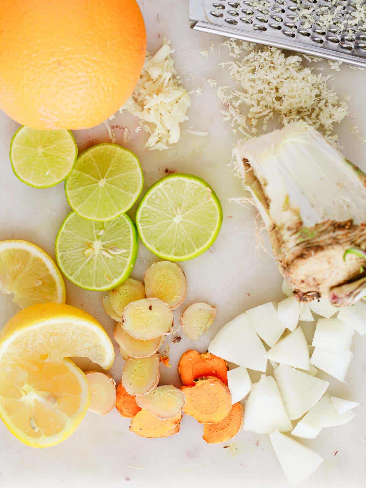 Sliced lime, lemon, ginger, turmeric, onion, orange, and grated horseradish on a white cutting board.