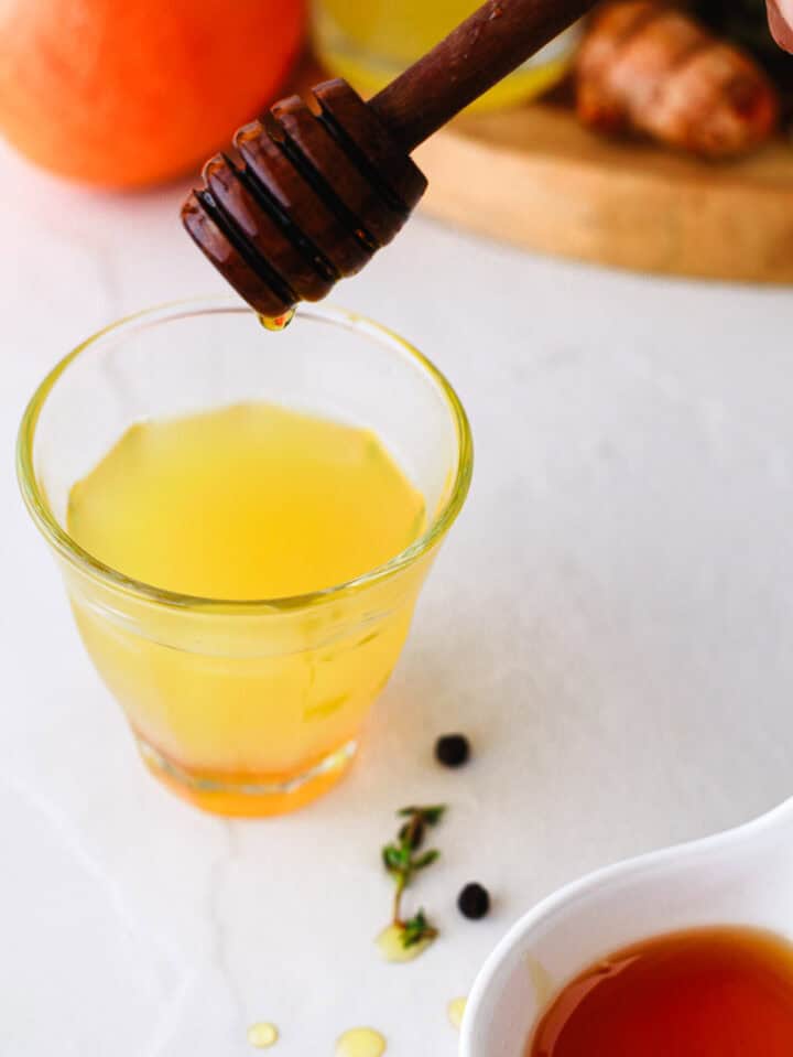 A honey dipper drips honey into a glass of yellow liquid, with herbs and peppercorns nearby.