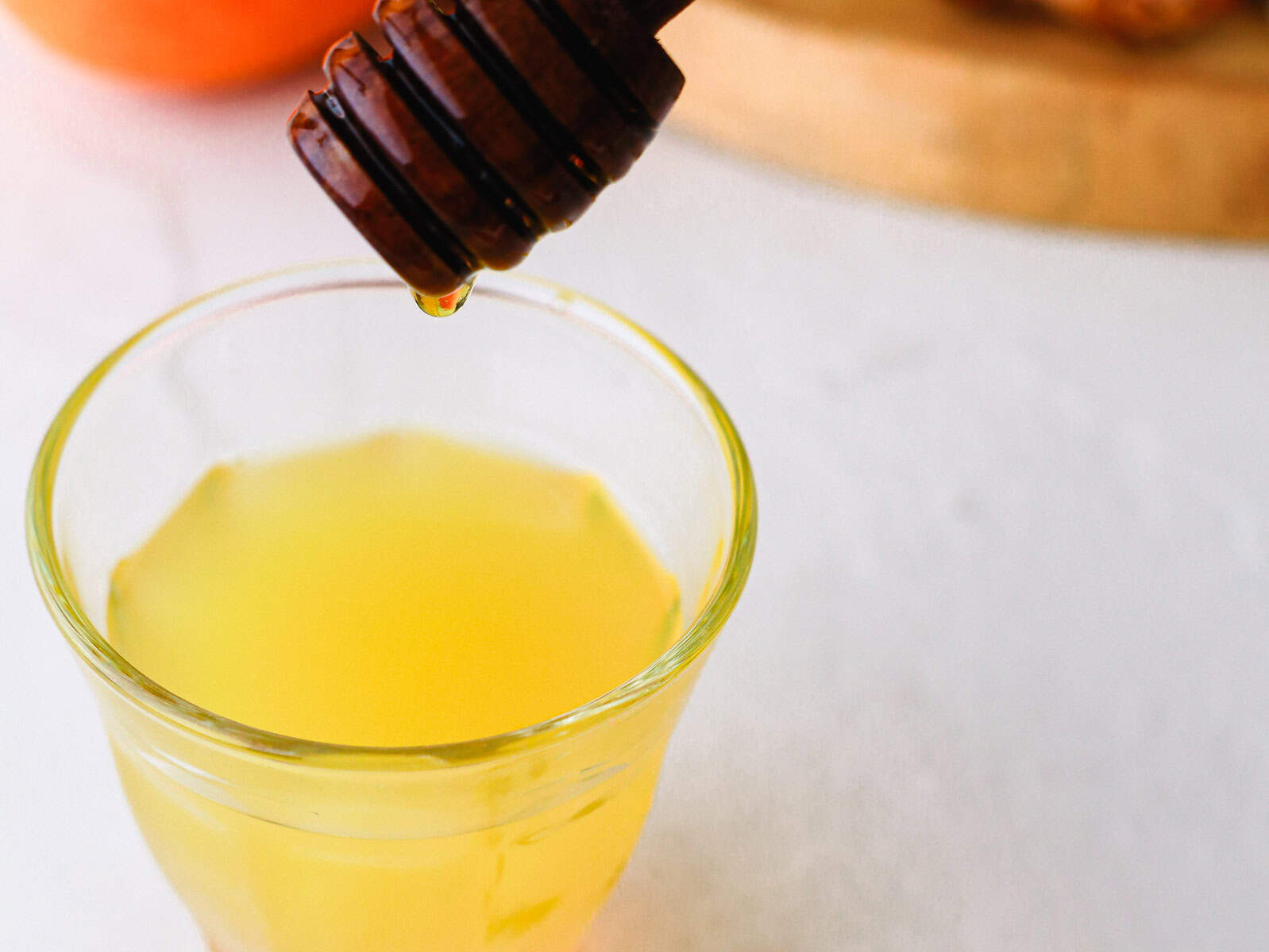 A honey dipper drizzling honey into a glass of yellow liquid on a white surface.
