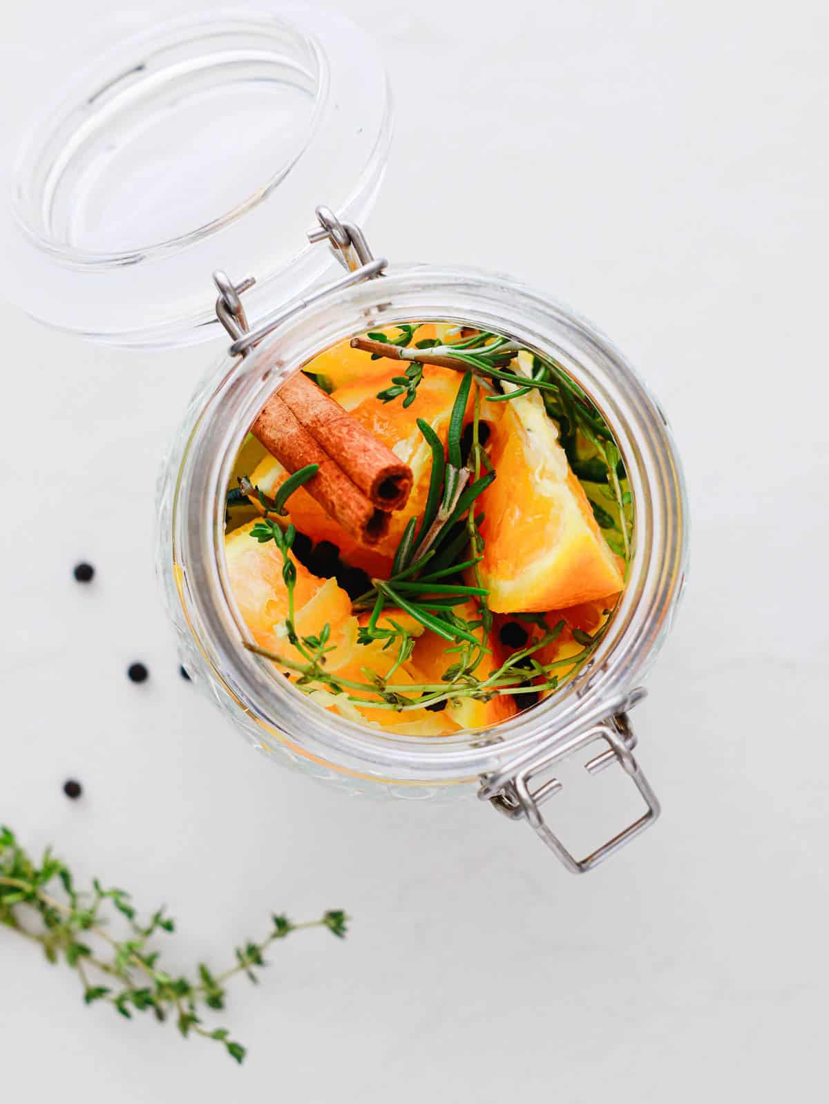 Open glass jar with orange slices, cinnamon stick, fresh herbs, and peppercorns on a white surface.