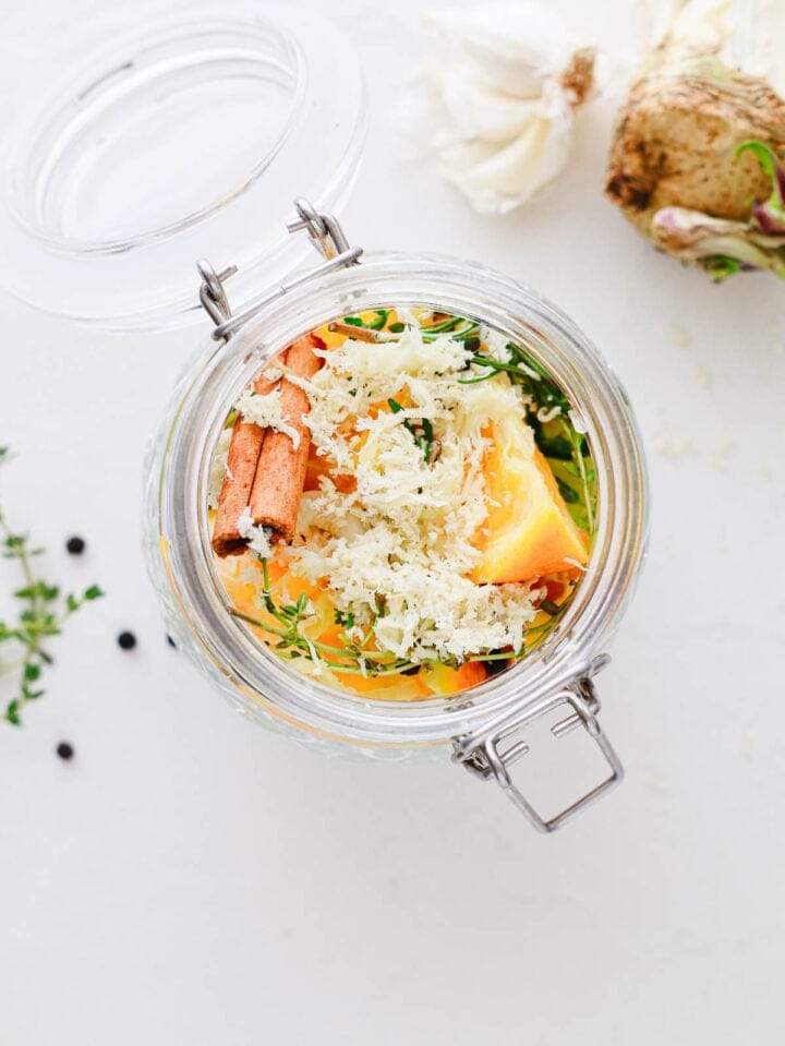 An open glass jar with grated horseradish, orange slices, cinnamon stick, and fresh herbs inside.