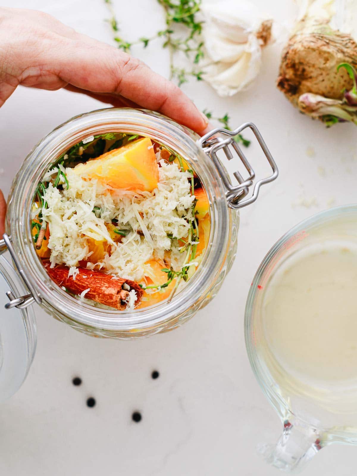 A hand holds a jar filled with vegetables, sauerkraut, and herbs next to a measuring cup of liquid.
