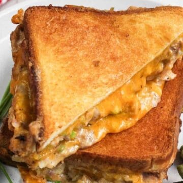 Grilled cheese sandwich with melted filling on a plate, garnished with parsley and pickles.