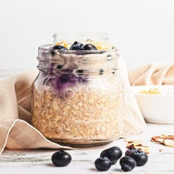 A jar of Blueberry Coconut Almond Overnight Oats topped with blueberries and almond slices sits on a cloth with scattered blueberries.