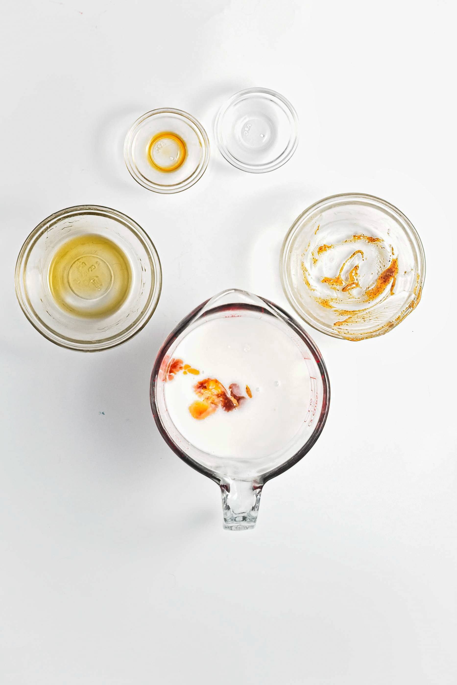 Five small glass bowls with various liquids and spices for Blueberry Coconut Almond Overnight Oats are arranged on a white background, viewed from above.