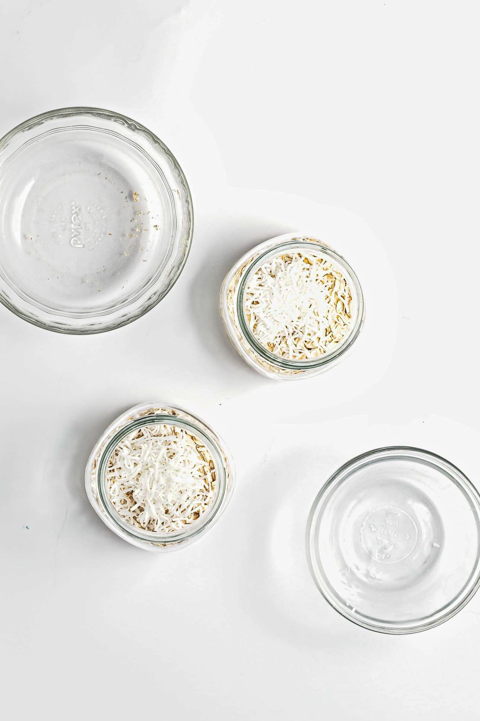 Three small glass jars with shredded cheese, two empty glass bowls, and a serving of Blueberry Coconut Almond Overnight Oats on a white surface.
