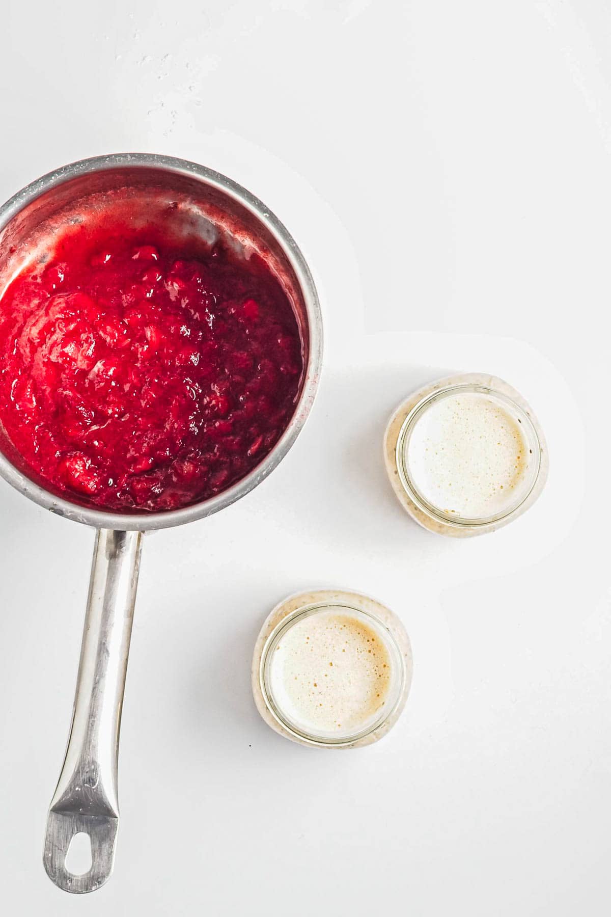 A saucepan with red berry sauce beside two small jars filled with a creamy, light-colored Strawberry Almond Overnight Chia Oats mixture.