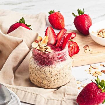 A jar of Strawberry Almond Overnight Chia Oats topped with strawberry slices and almond flakes, surrounded by fresh strawberries.