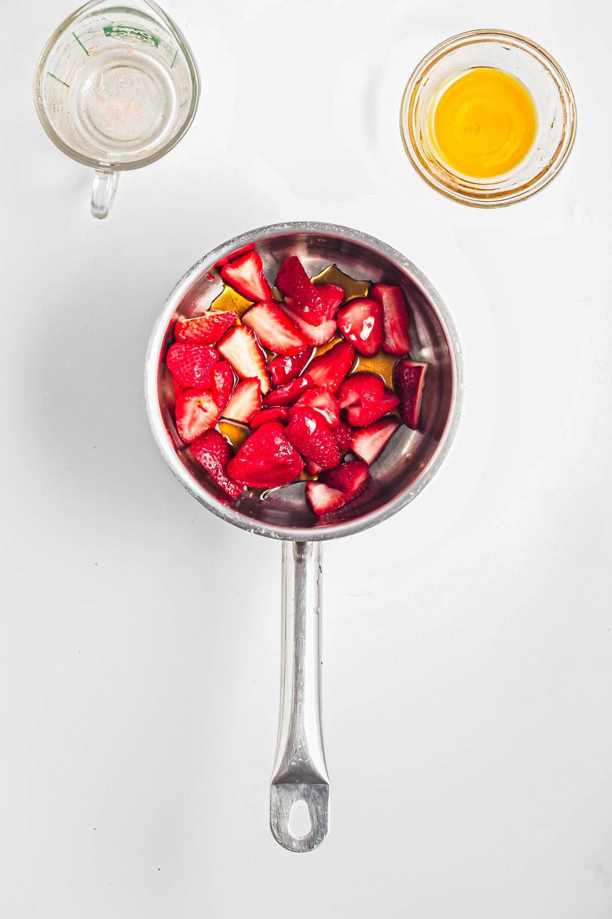 A saucepan with chopped strawberries, a measuring cup, and a small bowl of orange liquid on a white surface-perfect for preparing Strawberry Almond Overnight Chia Oats.