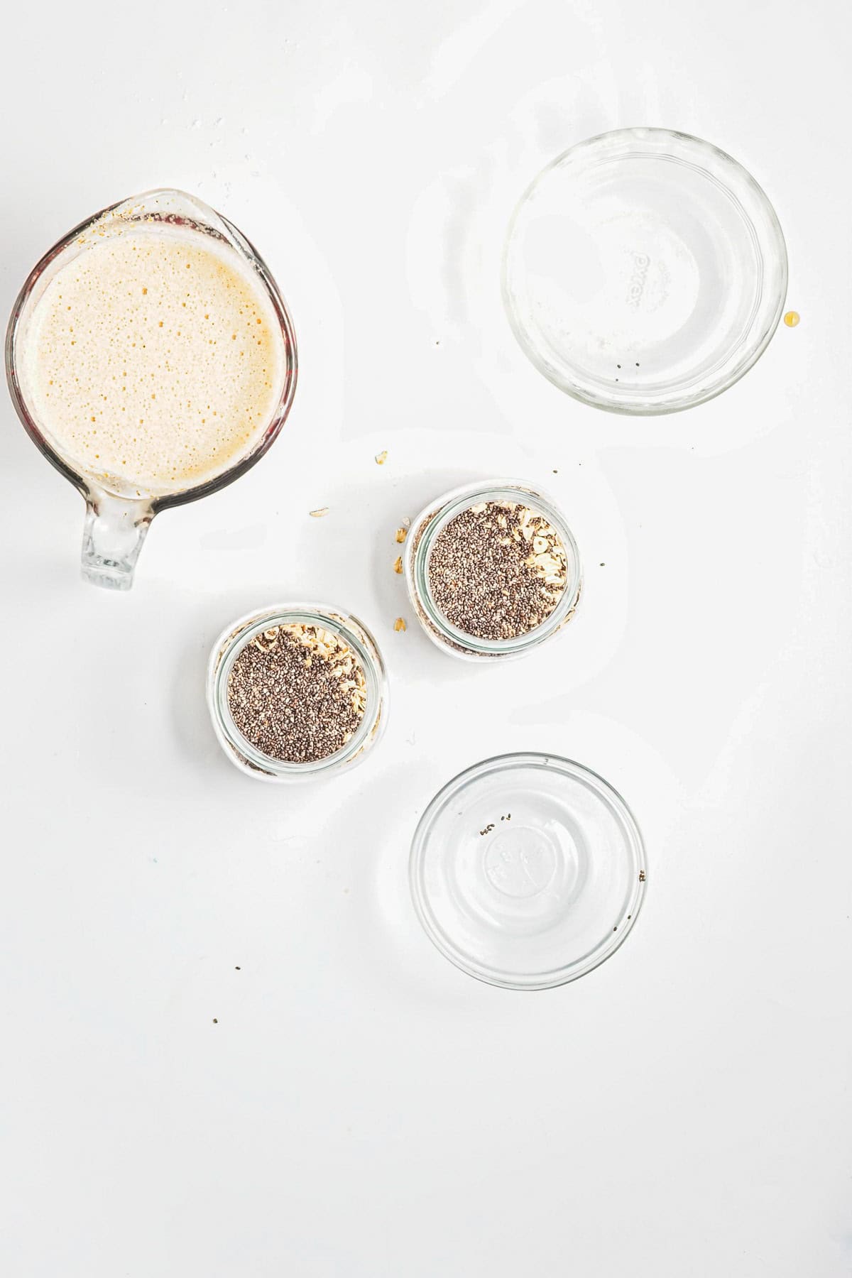 Overhead view of a measuring cup with creamy liquid and two jars of Strawberry Almond Overnight Chia Oats on a white surface.