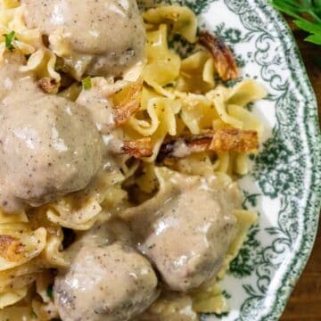 Meatballs with creamy gravy served over egg noodles on a decorative plate, garnished with fresh parsley.