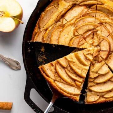 Skillet apple tart with several slices cut, next to a fork, plate, and halved apple on a white table.