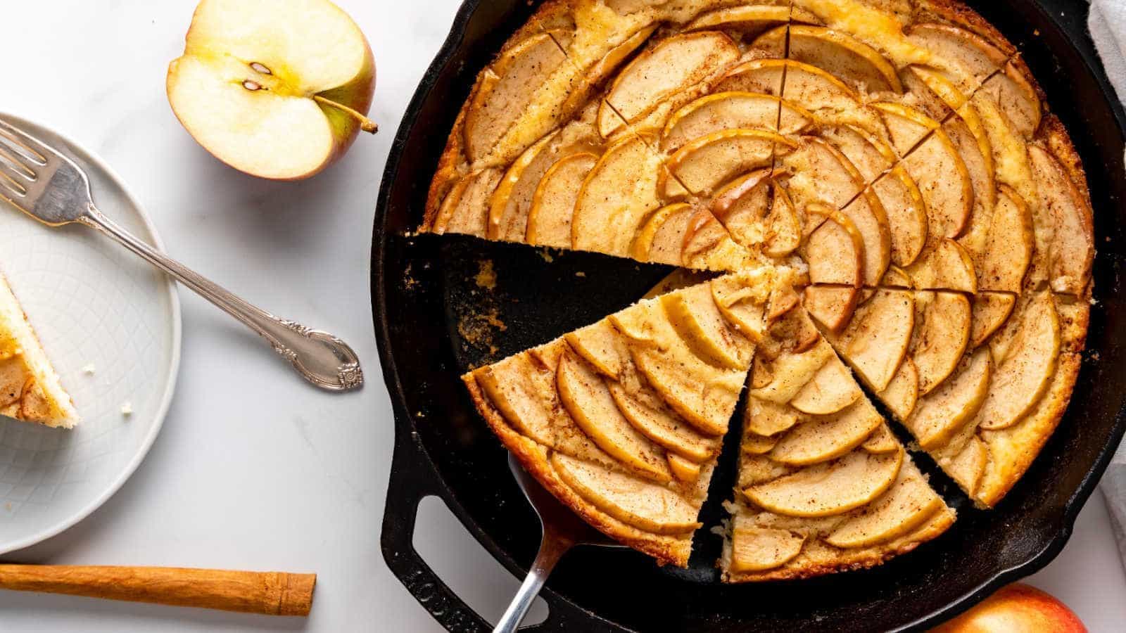 Skillet apple tart with several slices cut, next to a fork, plate, and halved apple on a white table.