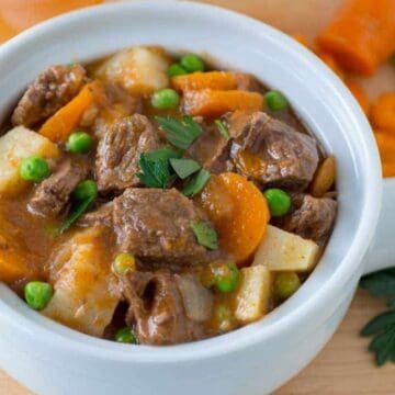 A bowl of beef stew with carrots, peas, potatoes, and fresh parsley on a wooden surface.