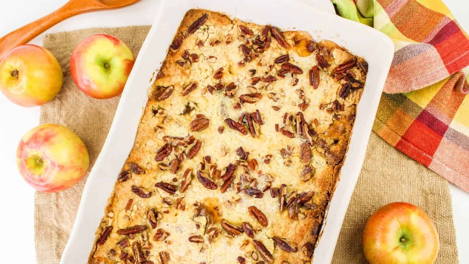 A baked apple dessert topped with pecans in a rectangular dish, surrounded by whole apples and a colorful napkin.