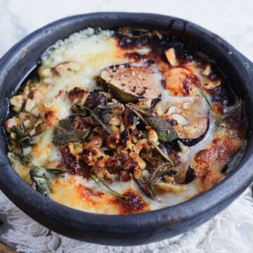 Baked cheese dish topped with herbs, nuts, and figs in a rustic black bowl.