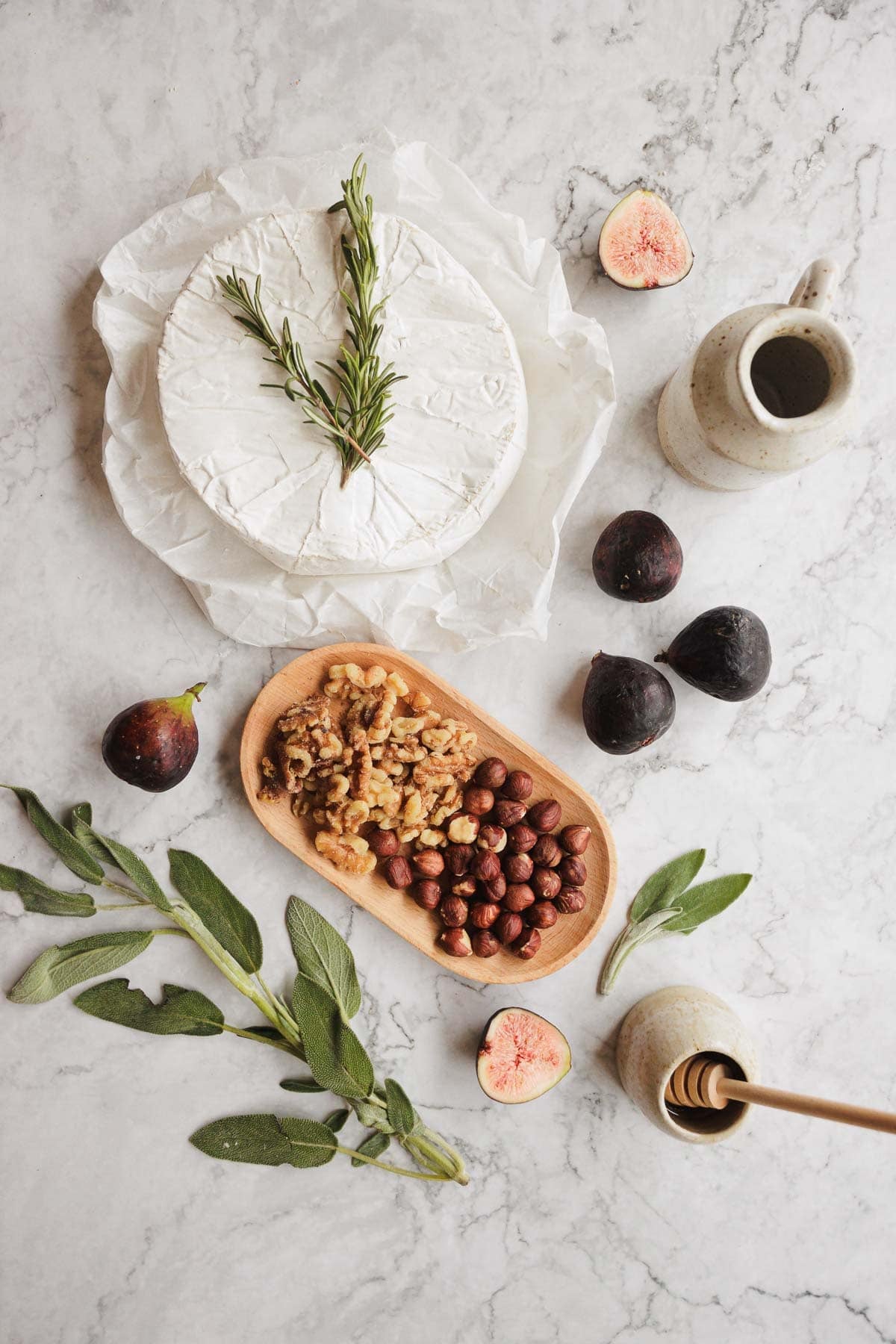 Round brie cheese with rosemary, figs, walnuts, almonds, honey dipper, and sage on a marble surface.