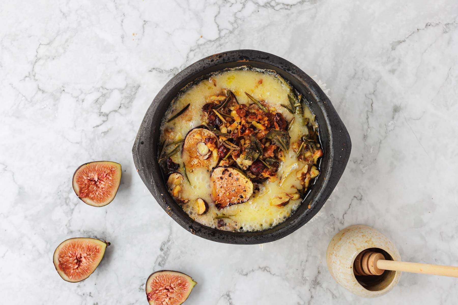 Baked cheese with vegetables in a black dish, figs, and a honey dipper on a marble surface.