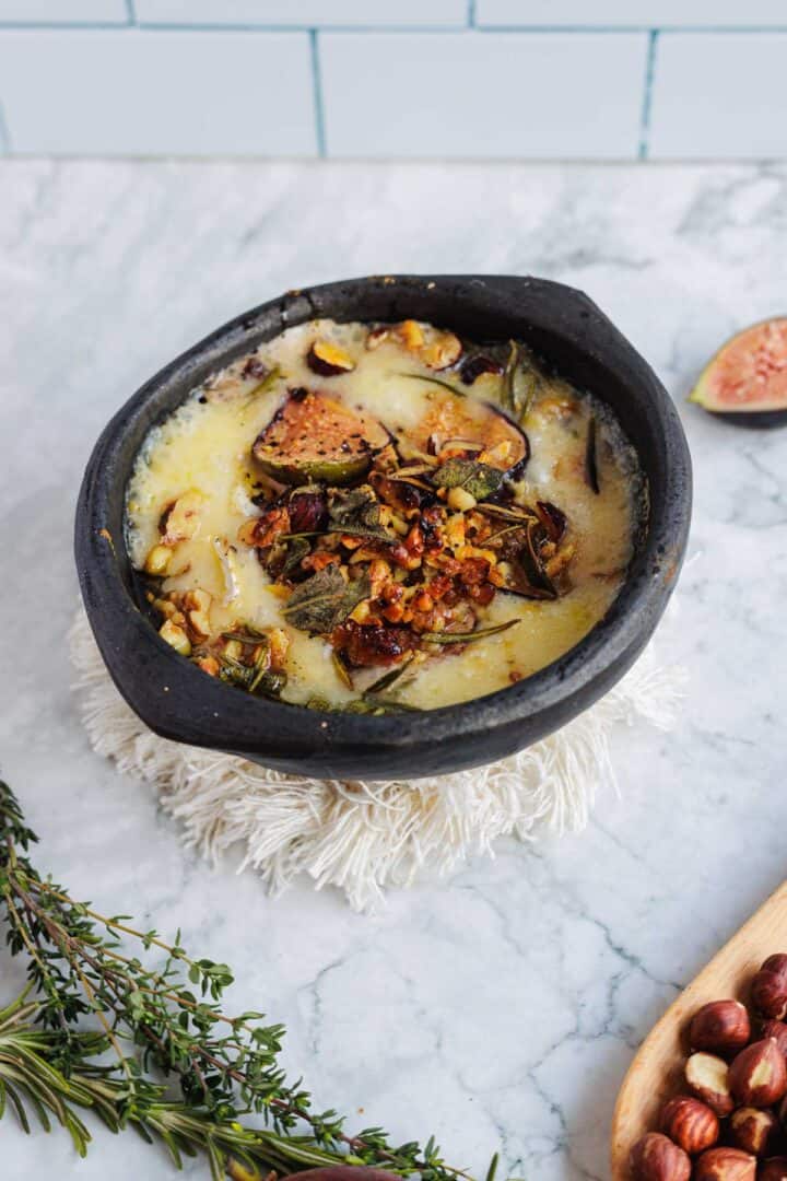 Baked brie topped with figs, herbs, and nuts in a black bowl on a marble countertop.