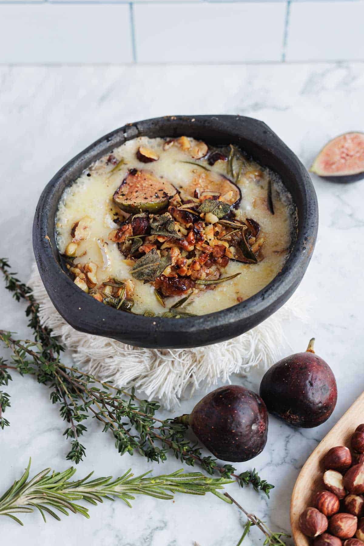 Baked brie with figs, nuts, and herbs in a black dish, with fresh figs and herbs on a marble surface.