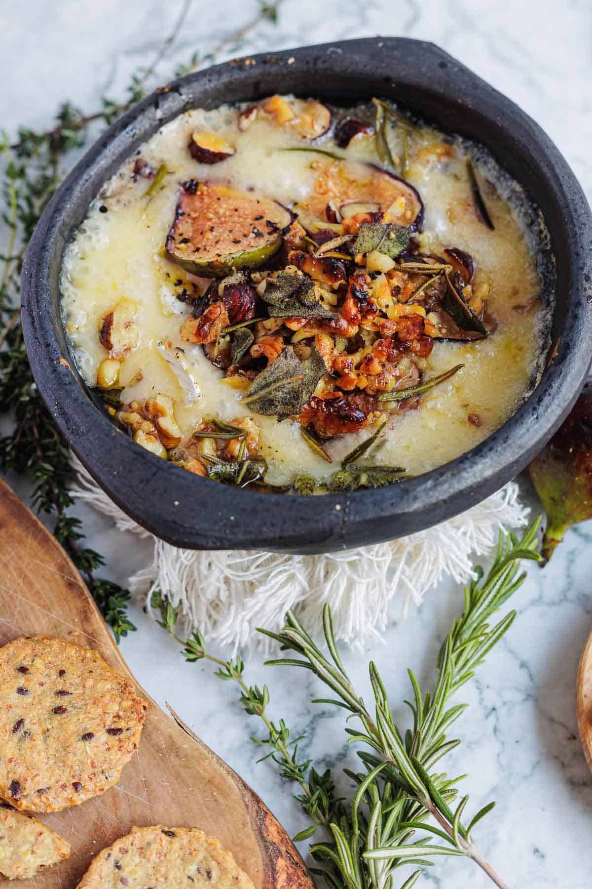 Baked cheese topped with herbs and nuts in a black bowl, surrounded by crackers and fresh rosemary.