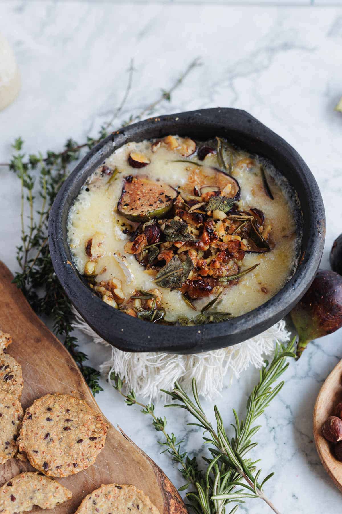 A black bowl of baked cheese topped with herbs, nuts, and figs, next to seeded crackers and fresh rosemary.