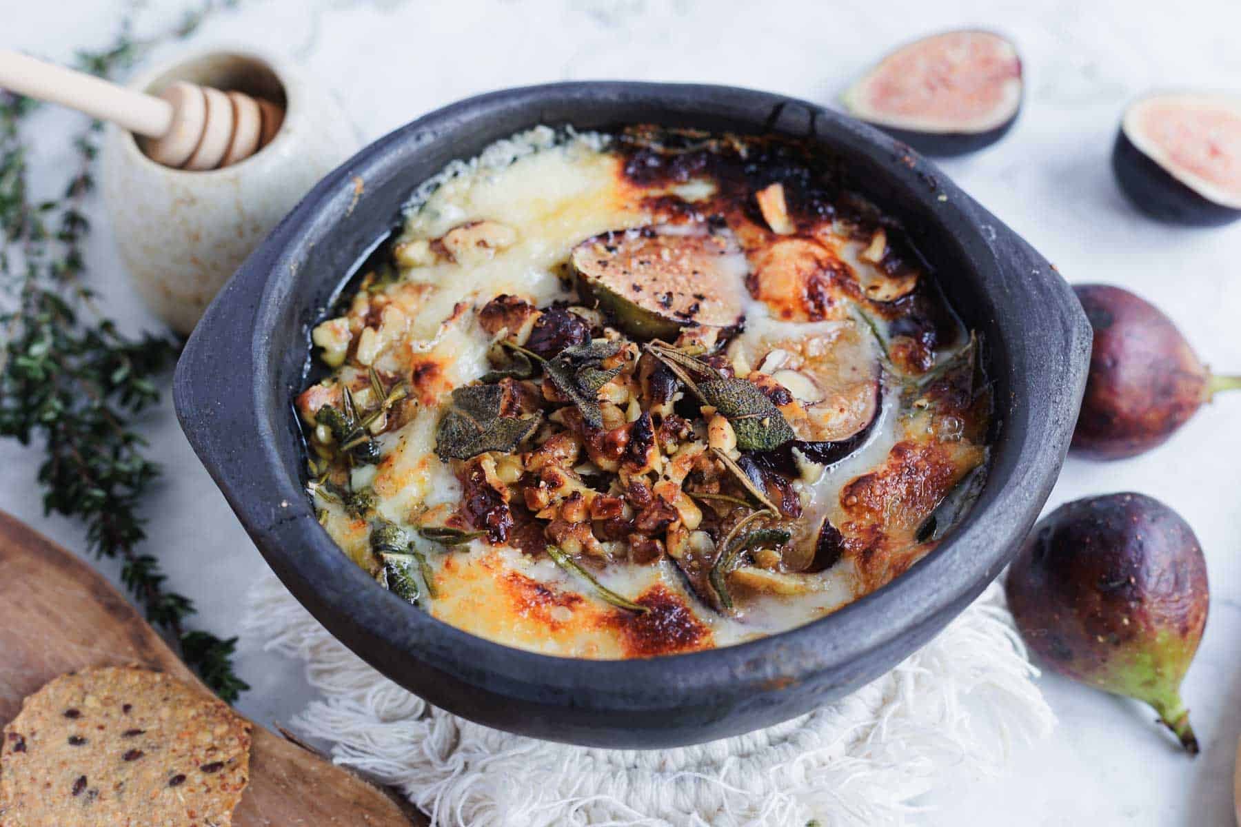 Baked cheese dip with figs, herbs, and walnuts in a black bowl, surrounded by fresh figs and honey.