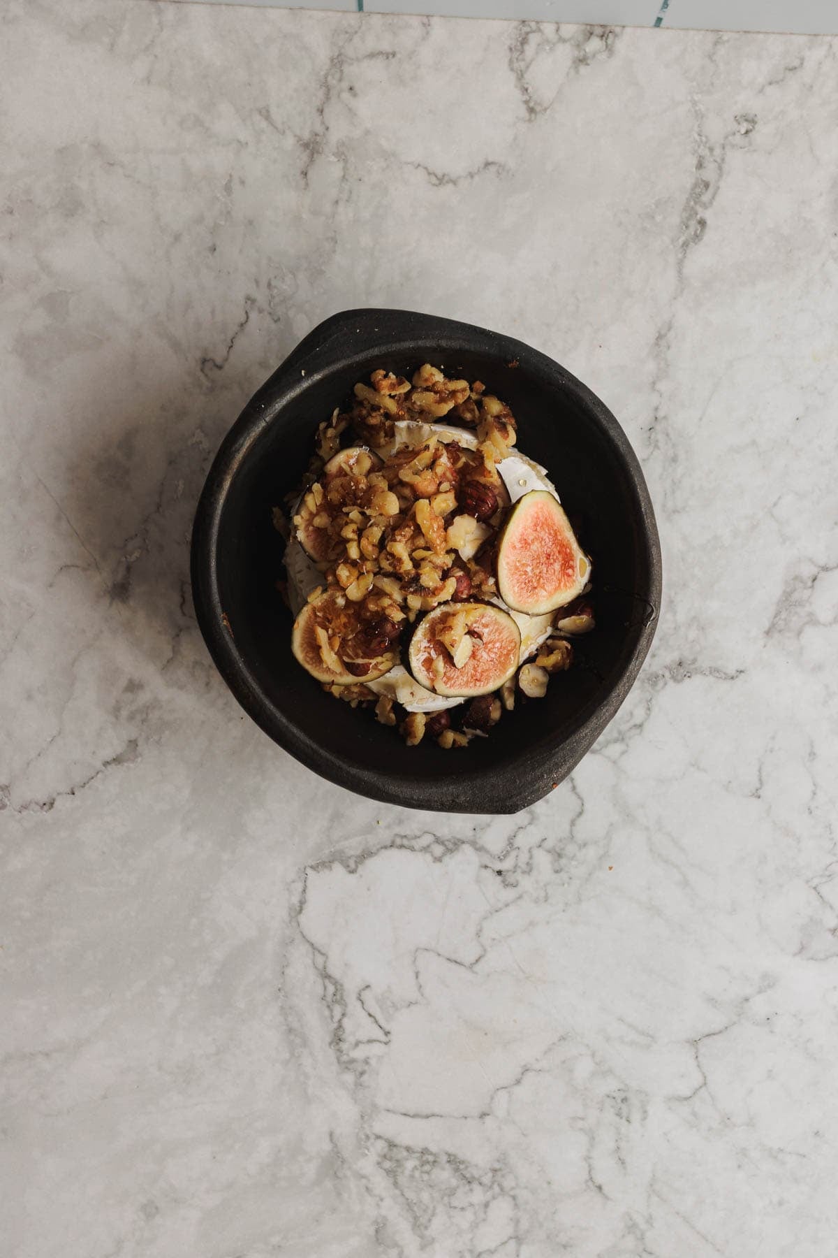 A black bowl with granola, figs, and walnuts on a light marble surface.