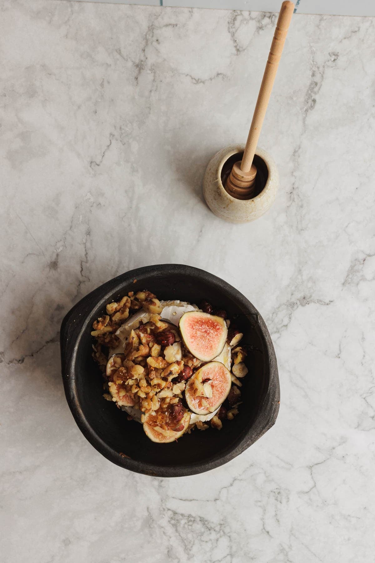 Black bowl with figs, walnuts, and cheese on marble surface; honey dipper in a small pot nearby.