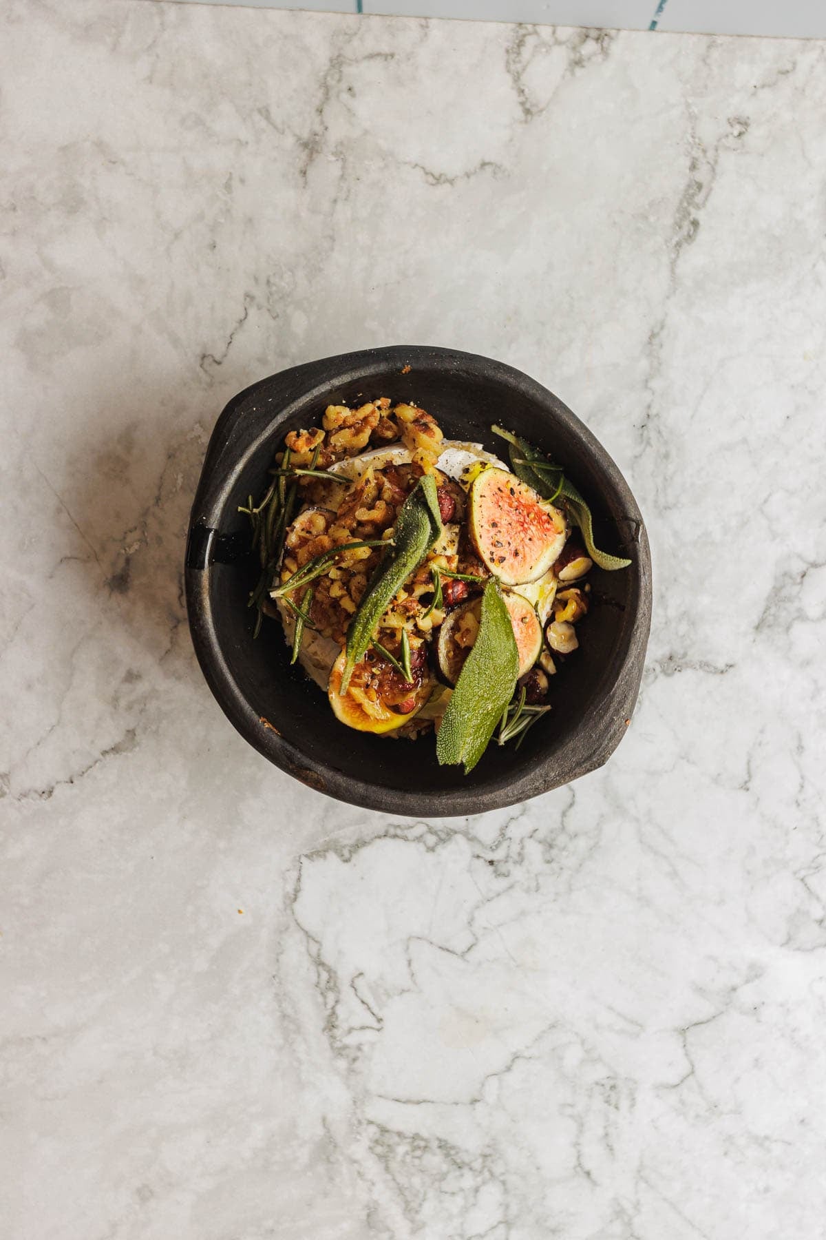 A black bowl with a salad of figs, greens, and grains on a marble surface.