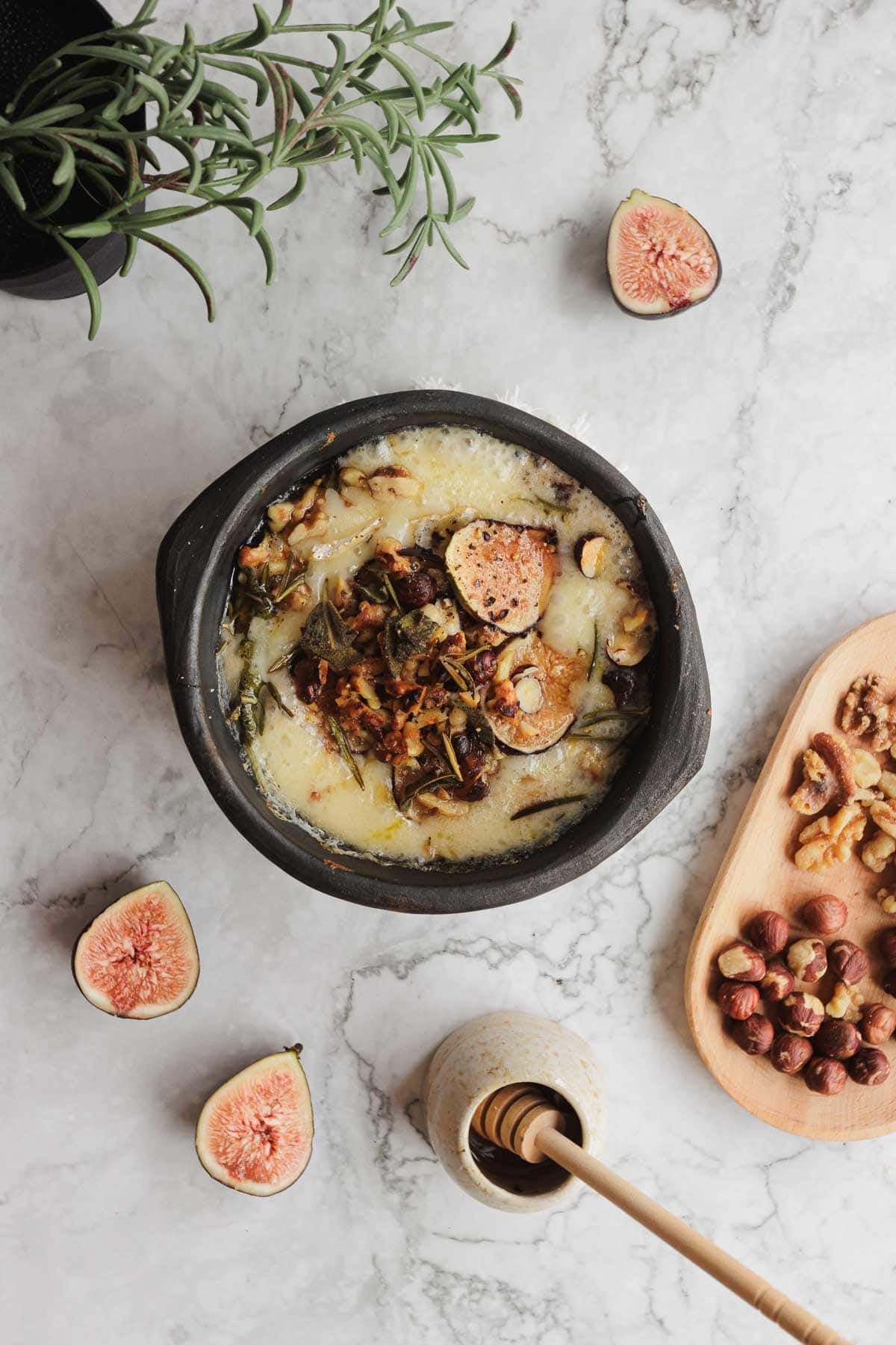 Top view of a baked cheese dish with figs, nuts, and herbs on a marble surface with honey and fresh figs nearby.