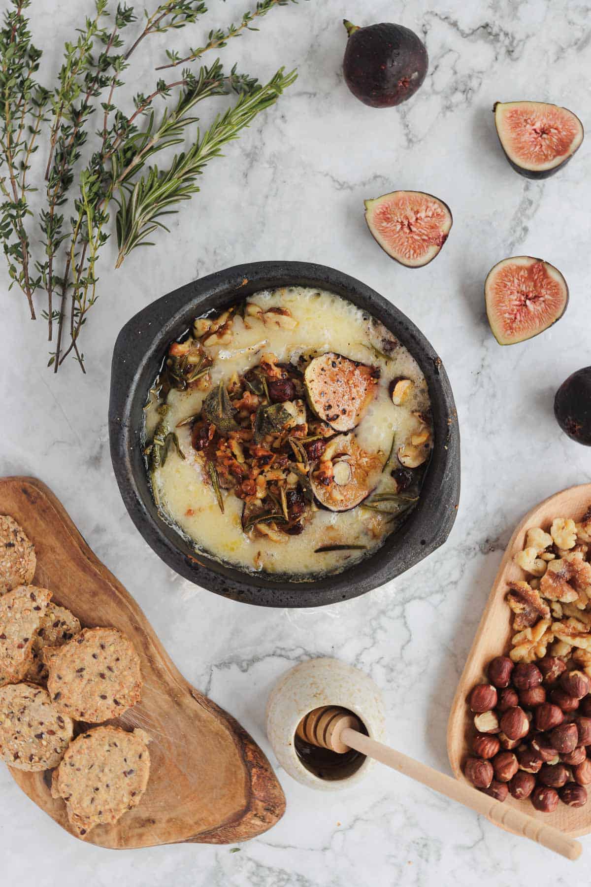 Baked cheese with herbs, figs, nuts, crackers, and honey on a marble surface.