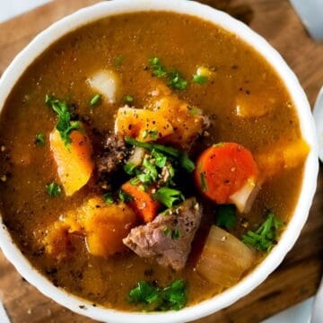 Bowl of beef and vegetable soup with carrots, potatoes, and herbs, on a wooden board beside a spoon.