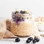 A jar of Blueberry Coconut Almond Overnight Oats topped with blueberries and sliced almonds, with a beige cloth and fresh blueberries nearby.