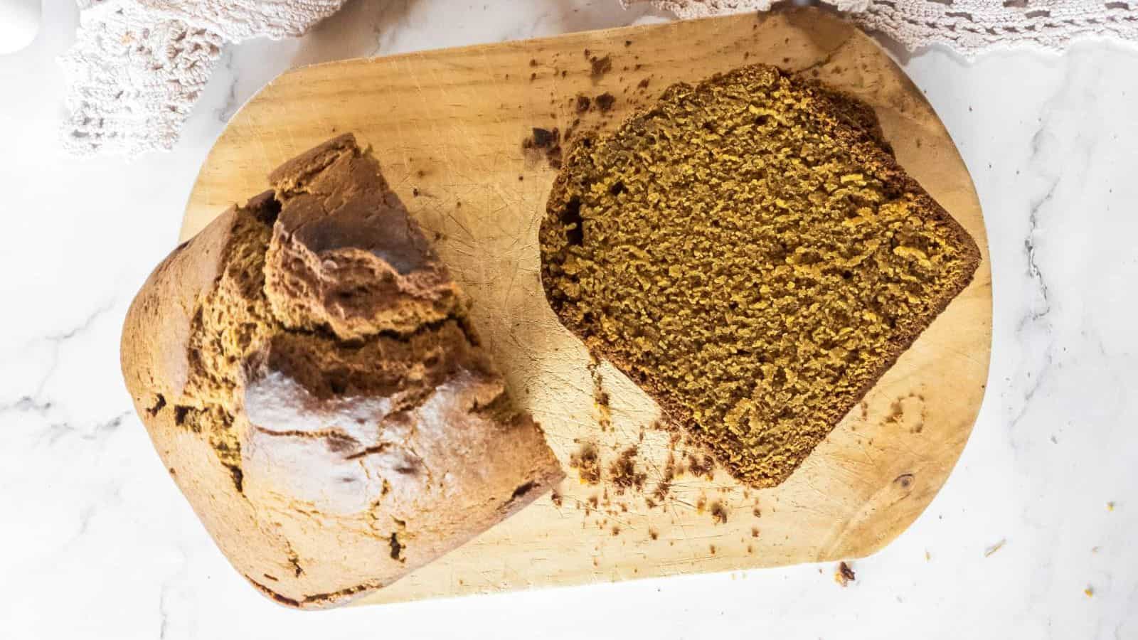 A slice of brown bread rests on a wooden cutting board next to the main loaf.