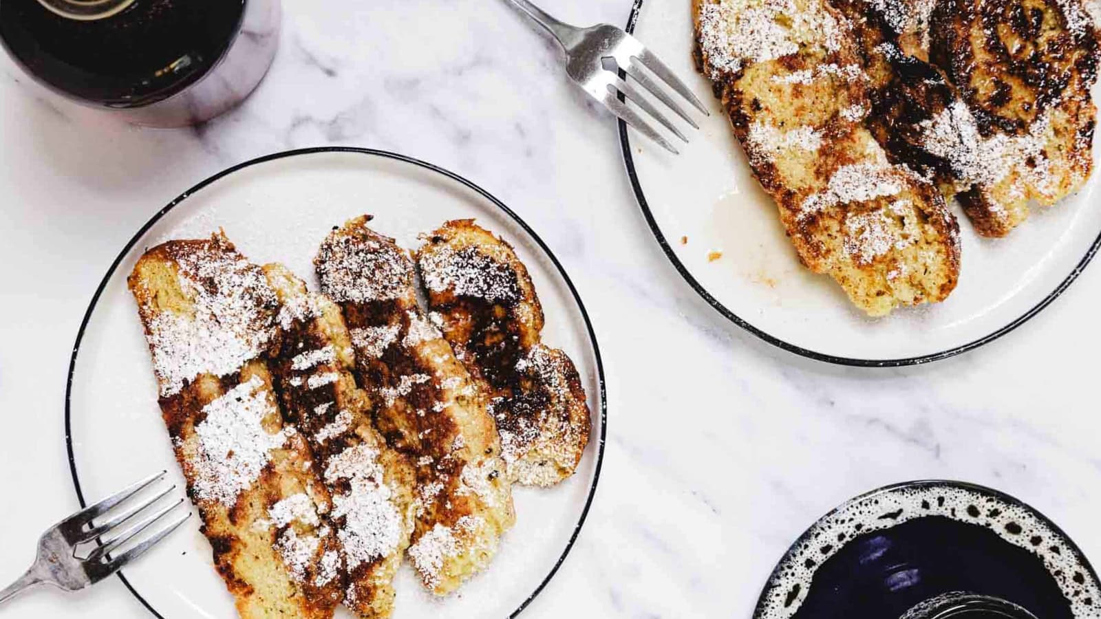 Four slices of French toast topped with powdered sugar on a white plate, with a fork beside them.