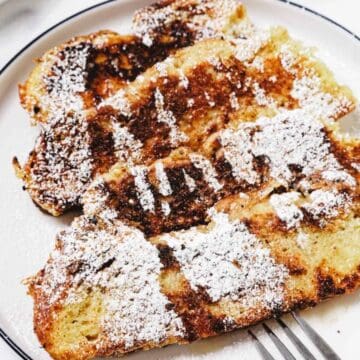 Three slices of French toast topped with powdered sugar on a white plate, with a fork beside them.