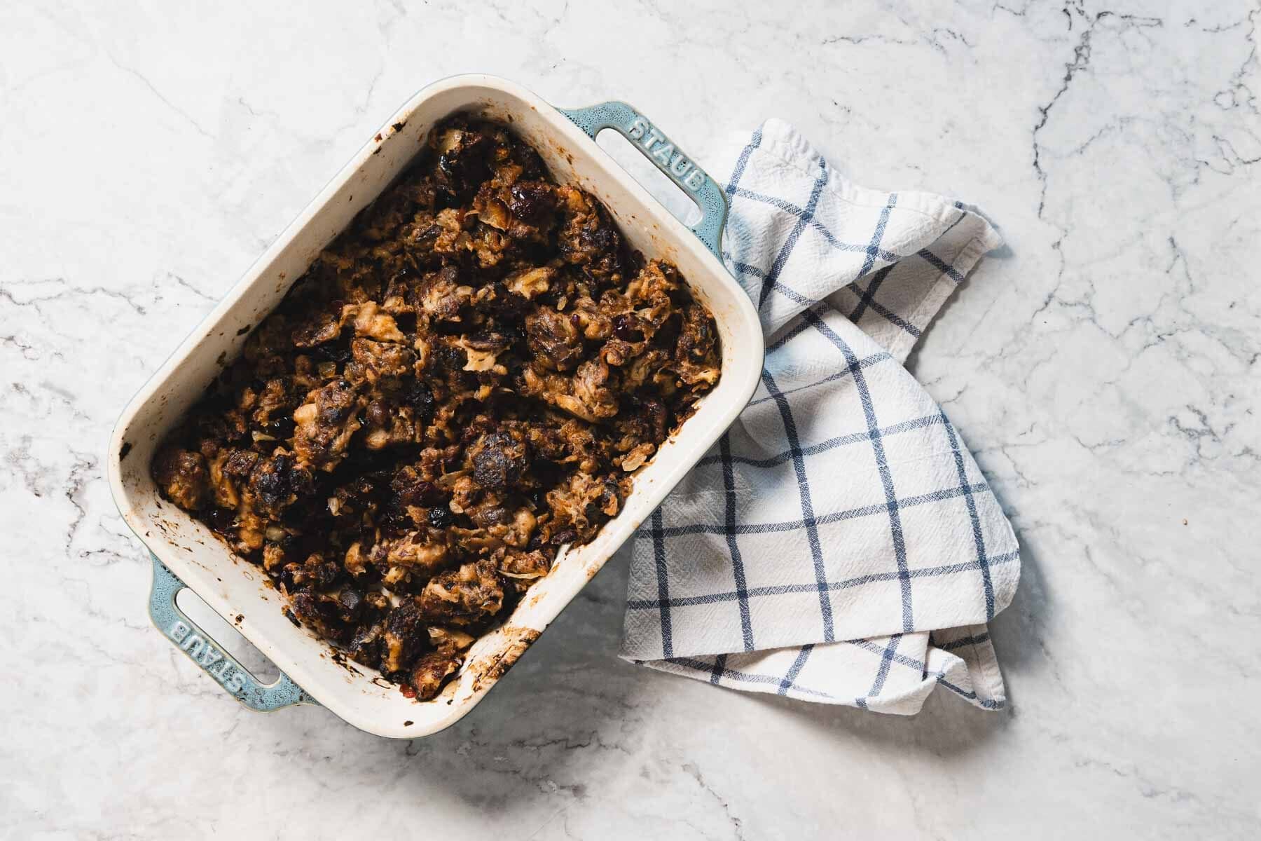 A baking dish with browned gluten free stuffing sits next to a folded blue and white checkered kitchen towel on marble.