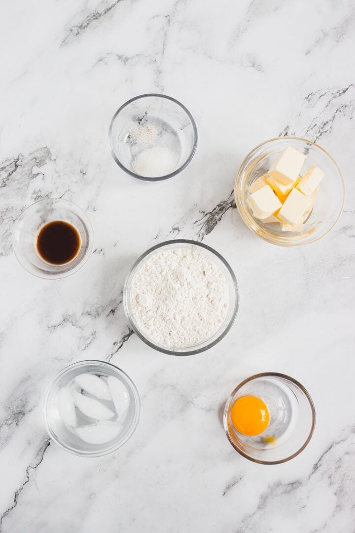 Six glass bowls with flour, egg yolk, butter, vanilla, water with ice, and a white powder on a marble surface.
