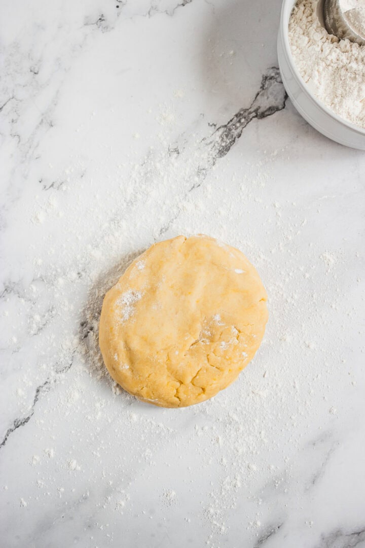 Round disc of dough on a floured marble surface, with a bowl of flour in the corner.