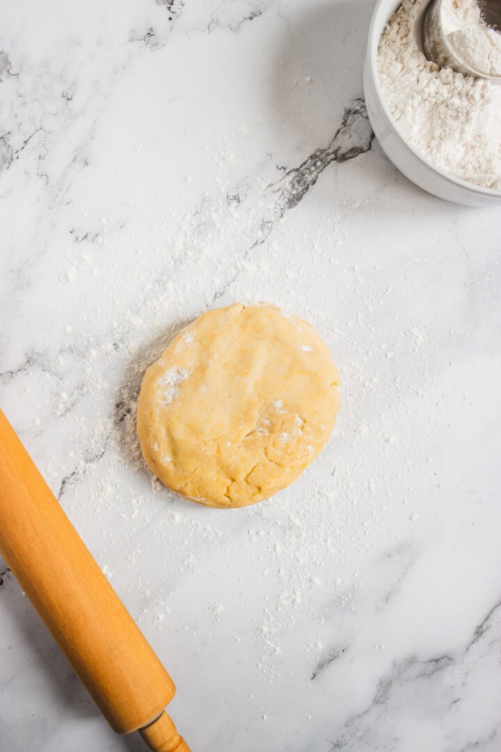 A ball of dough on a floured marble surface with a rolling pin and a bowl of flour nearby.