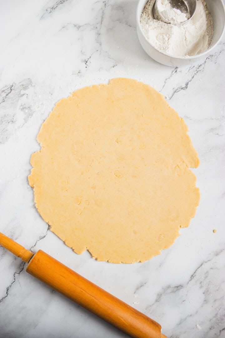 Rolled out dough on a marble surface with a wooden rolling pin and a bowl of flour nearby.