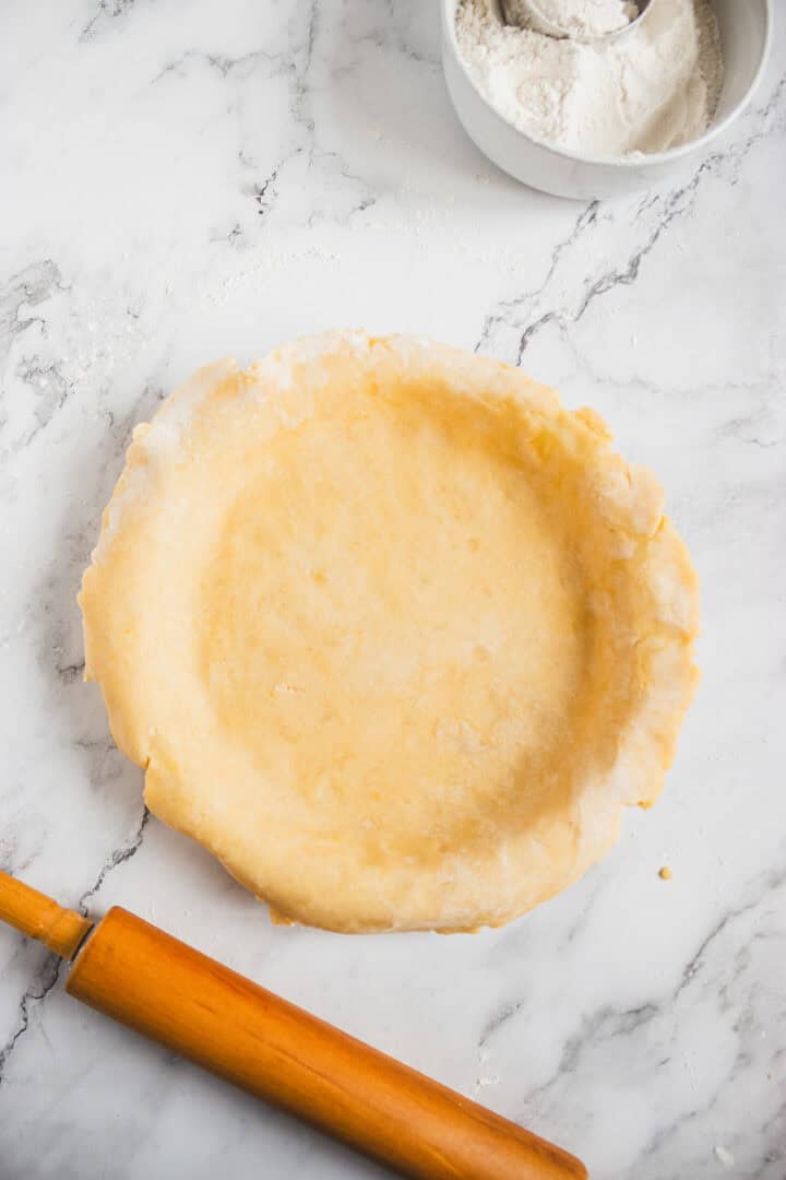 Unbaked pie crust in a pie dish, with a rolling pin and a bowl of flour on a marble surface.