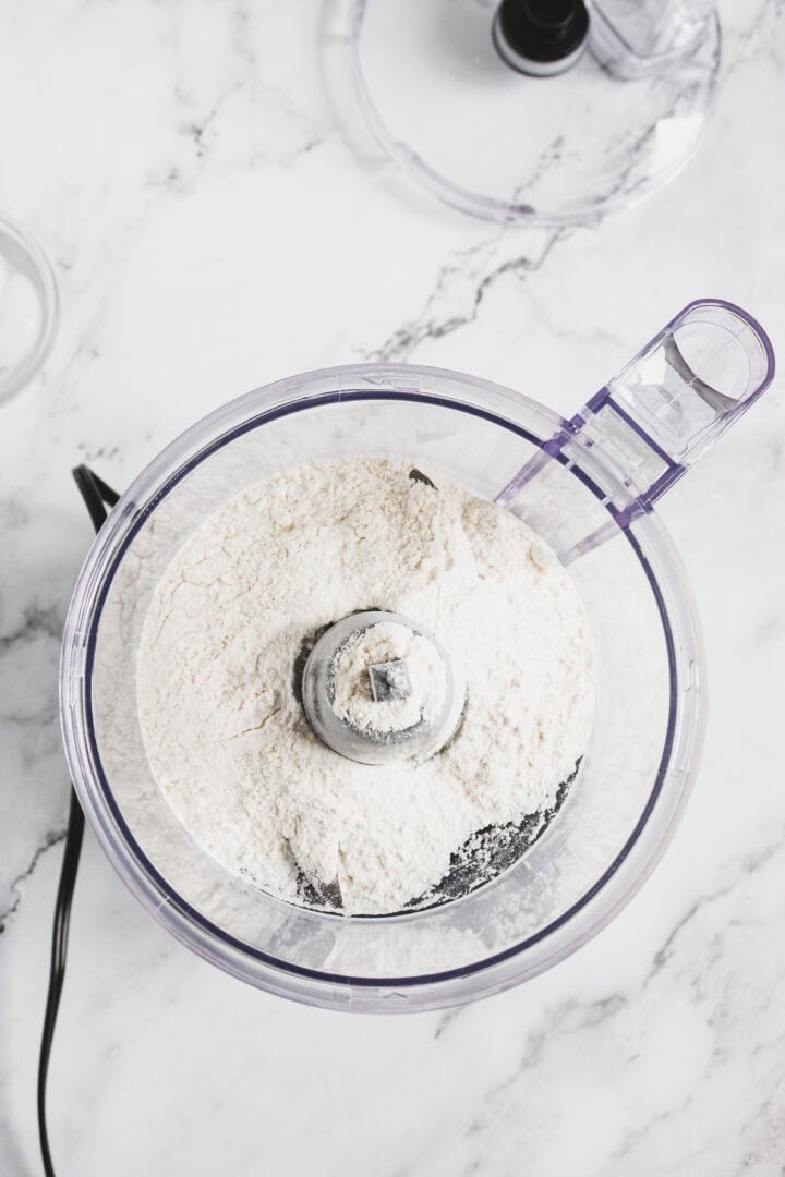 Overhead view of flour in a food processor bowl on a white marble countertop.