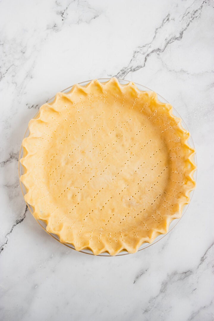 Unbaked pie crust with crimped edges and fork holes in a glass dish on a marble surface.