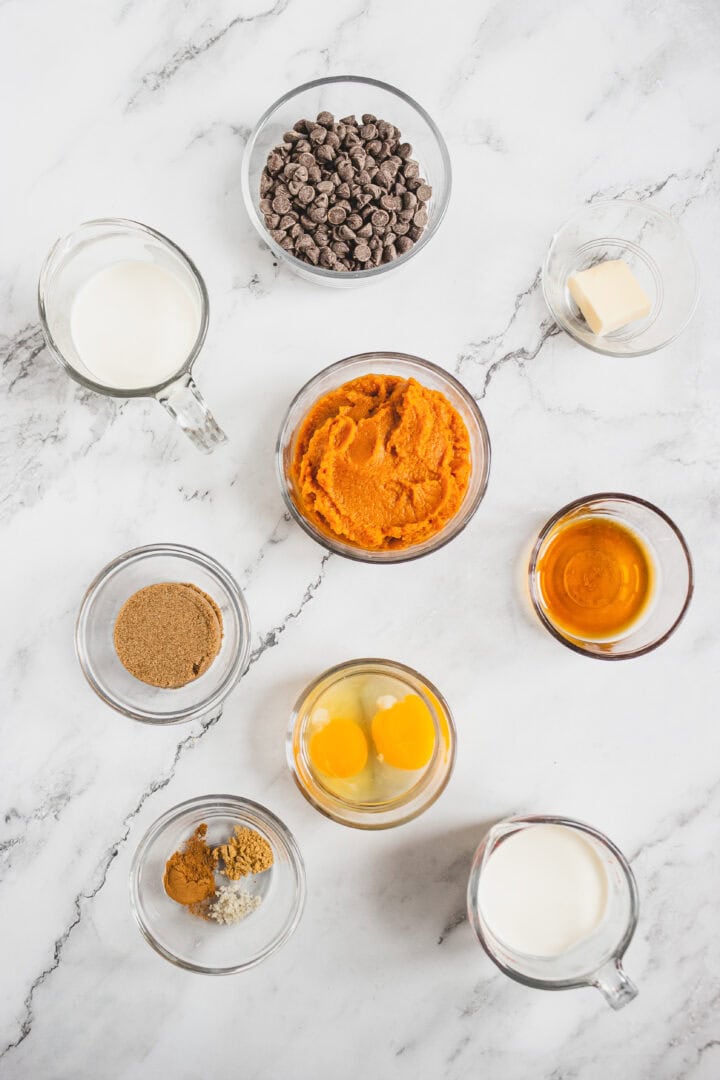 Baking ingredients arranged on a marble surface, including eggs, pumpkin puree, chocolate chips, and spices.