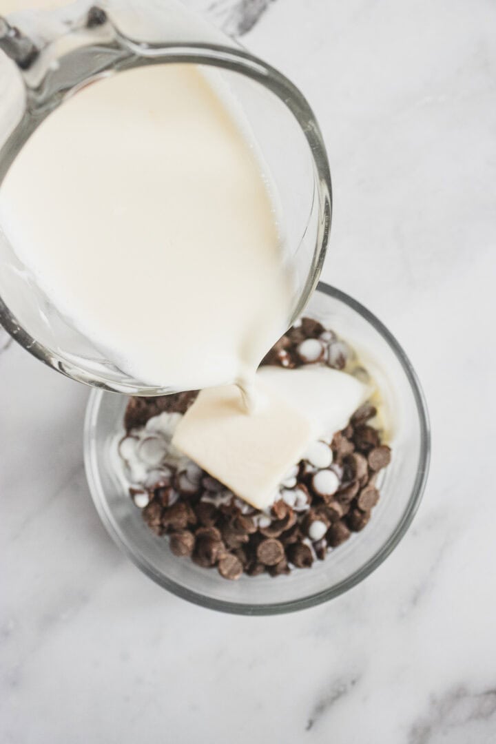 Cream being poured into a bowl of chocolate chips and butter on a white marble surface.