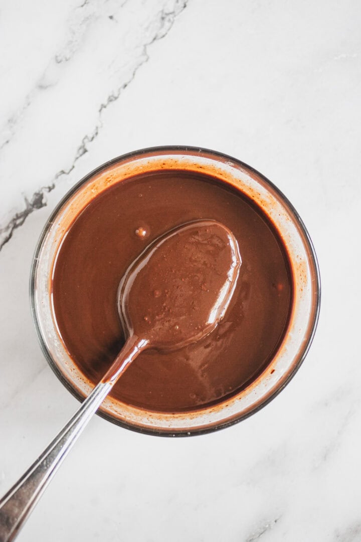 Bowl of smooth chocolate sauce with a spoon on a white marble surface.