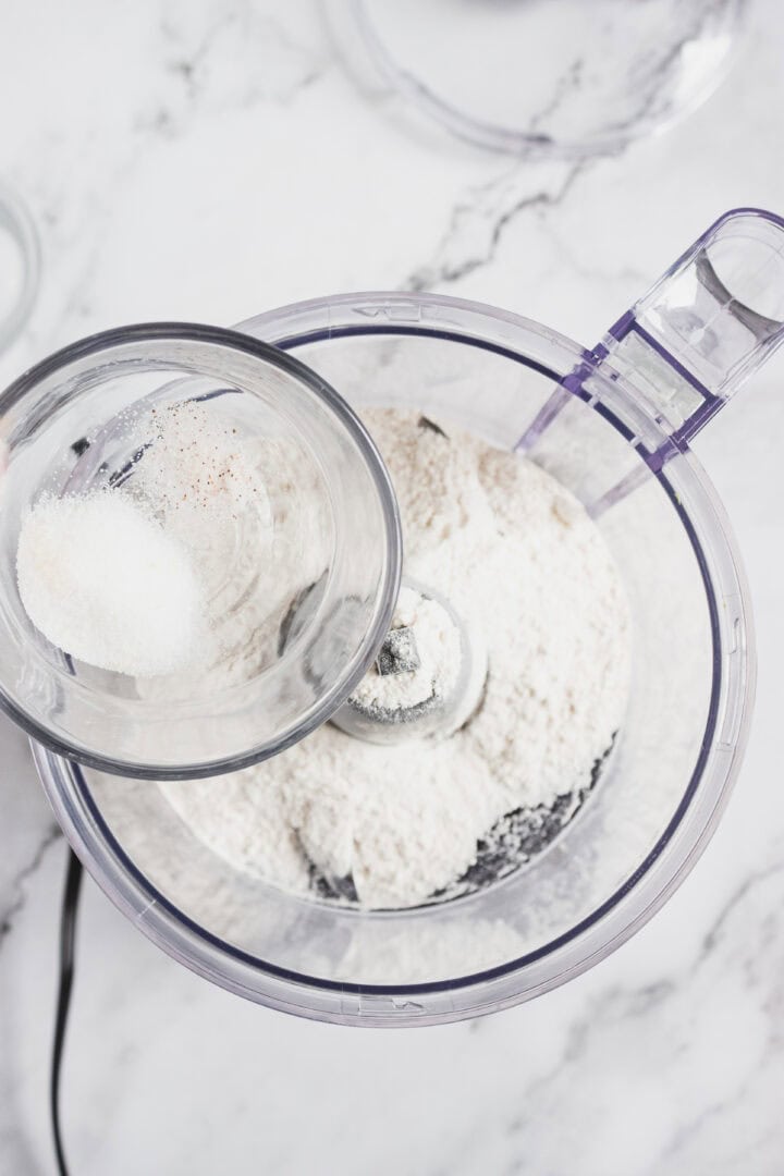 A glass cup with salt is being poured into a food processor filled with flour on a marble countertop.