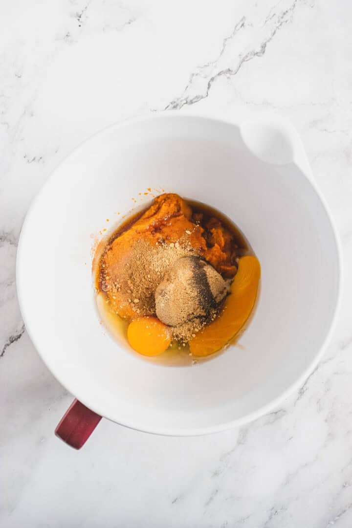 White bowl with eggs, pumpkin puree, sugar, and spices on a white marble surface.